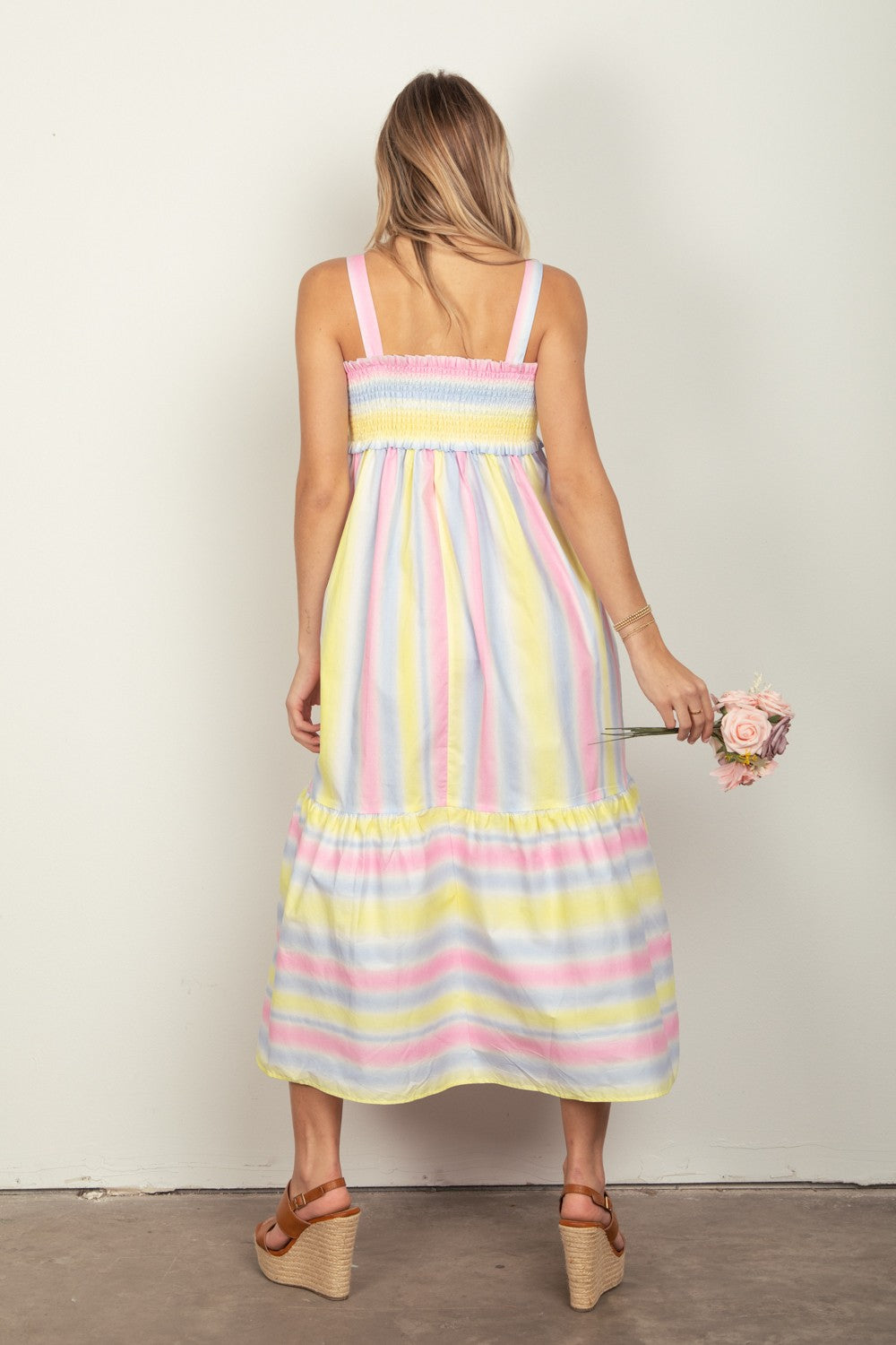 A woman with long hair, viewed from behind, wears a VERY J Striped Woven Smocked Midi Cami Dress. She holds a pink rose before a plain white wall and pairs the dress perfectly with brown wedge sandals.