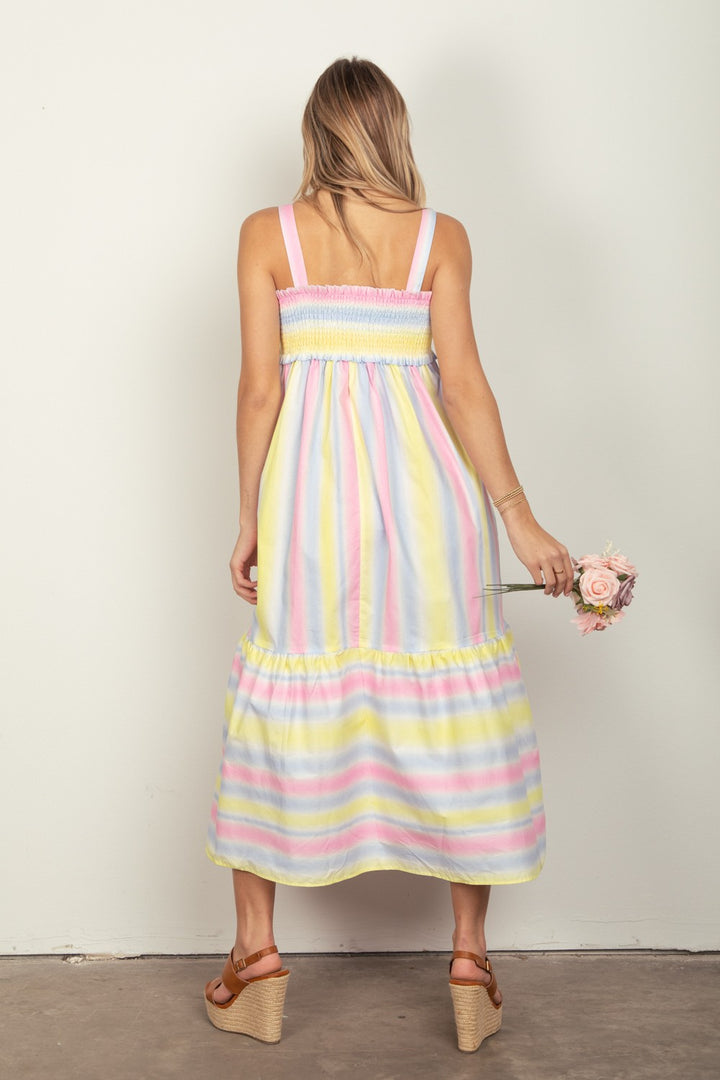 A woman with long hair, viewed from behind, wears a VERY J Striped Woven Smocked Midi Cami Dress. She holds a pink rose before a plain white wall and pairs the dress perfectly with brown wedge sandals.