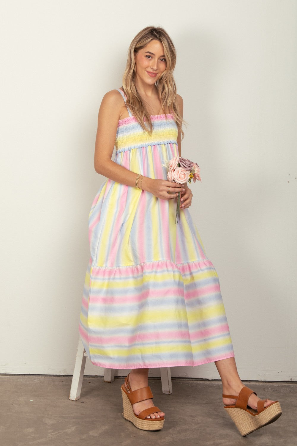 A woman with long blonde hair sits smiling on a white stool, wearing the VERY J Striped Woven Smocked Midi Cami Dress, holding pink roses. Her brown wedge sandals complement the look against an off-white wall.