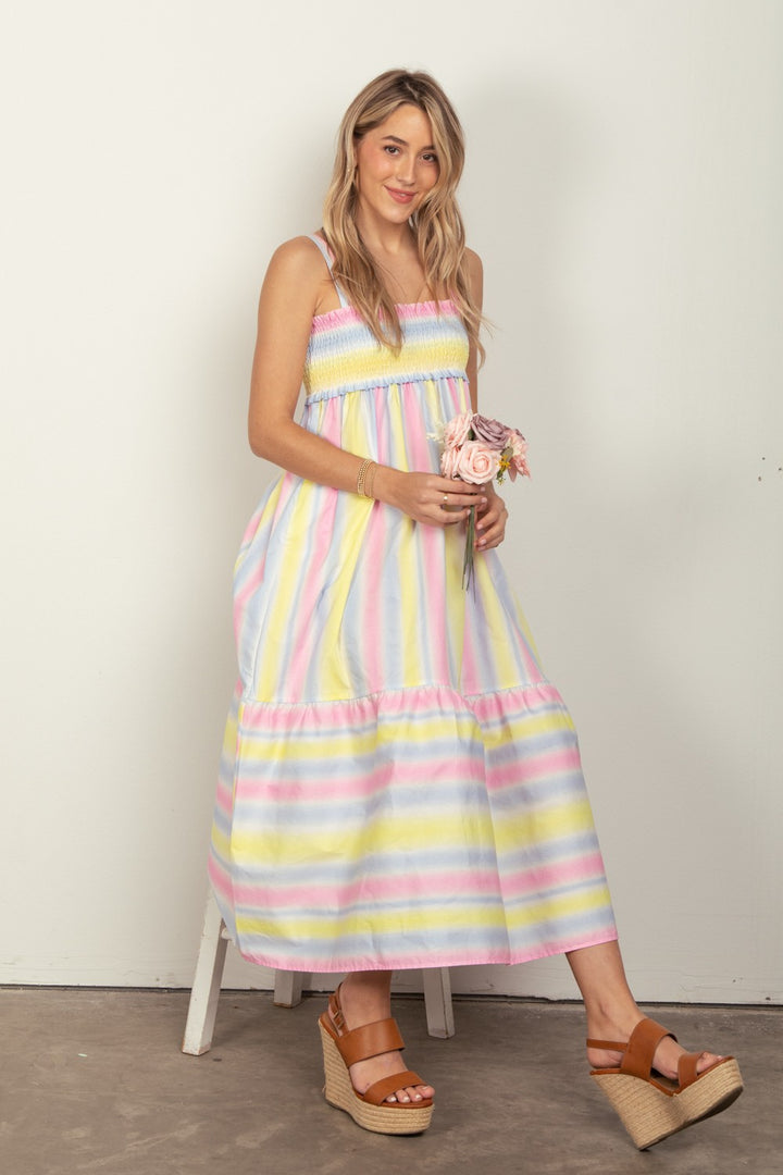 A woman with long blonde hair sits smiling on a white stool, wearing the VERY J Striped Woven Smocked Midi Cami Dress, holding pink roses. Her brown wedge sandals complement the look against an off-white wall.