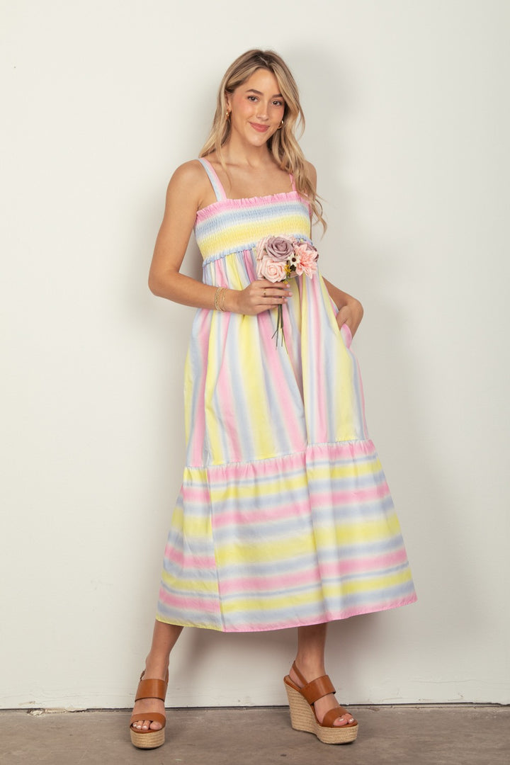 A woman with long blonde hair smiles while holding a small bouquet of pink roses, wearing the VERY J Striped Woven Smocked Midi Cami Dress and wedge sandals, standing against a plain background.