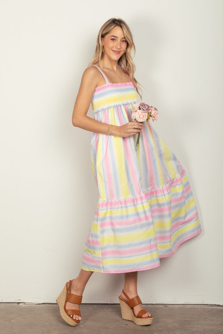 In front of a plain pale wall, a woman with light hair smiles while holding flowers. She wears wedge sandals and the VERY J Striped Woven Smocked Midi Cami Dress, featuring pastel-colored stripes, adjustable cami straps, a smocked bodice, sleeveless design, and ankle-length flow.