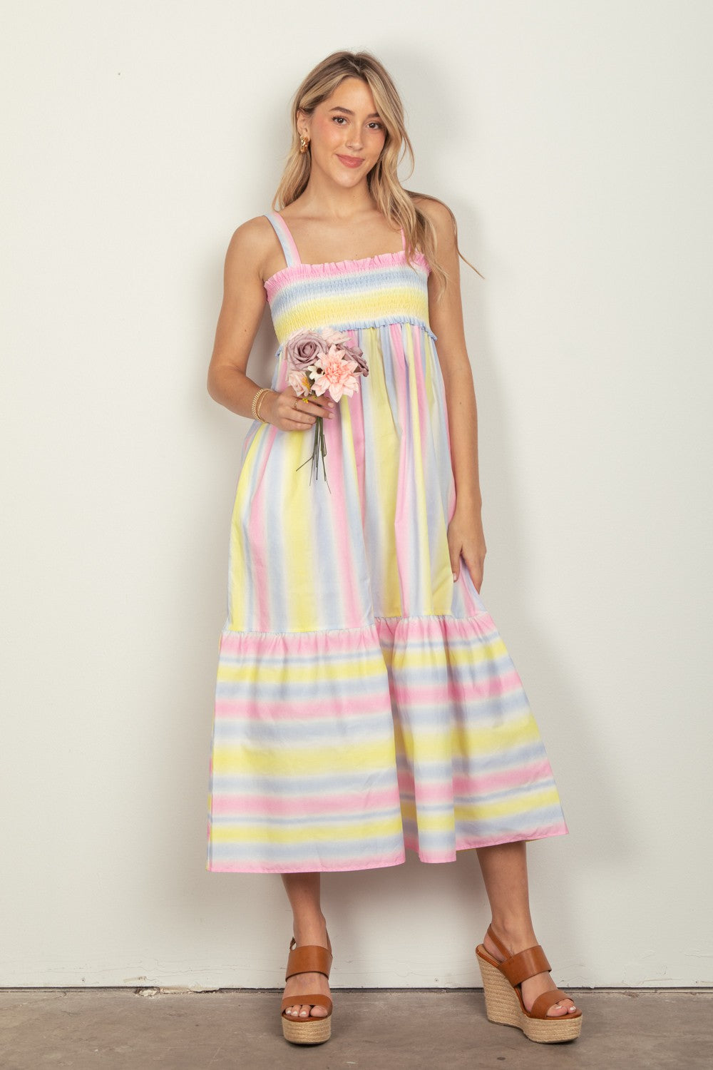 A woman in a VERY J Striped Woven Smocked Midi Cami Dress holds pink flowers, set against a light background. Tan platform sandals complement her look, and the dresss adjustable cami straps add elegance.