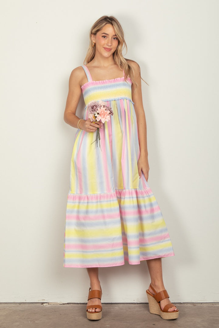 A woman in a VERY J Striped Woven Smocked Midi Cami Dress holds pink flowers, set against a light background. Tan platform sandals complement her look, and the dresss adjustable cami straps add elegance.