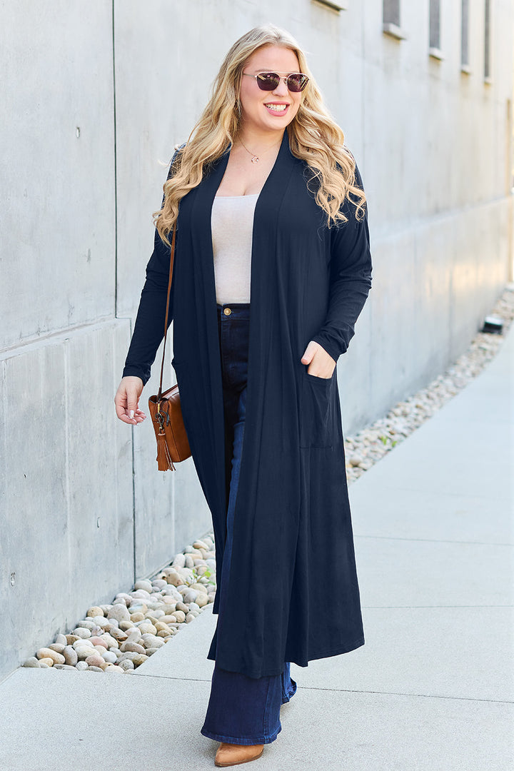 A woman with long blonde hair and sunglasses strolls on a sidewalk beside a pebble path. She wears the Basic Bae Full Size Open Front Long Sleeve Cover Up in black over a white top and dark jeans. With her brown shoulder bag, she smiles against the backdrop of a concrete wall.