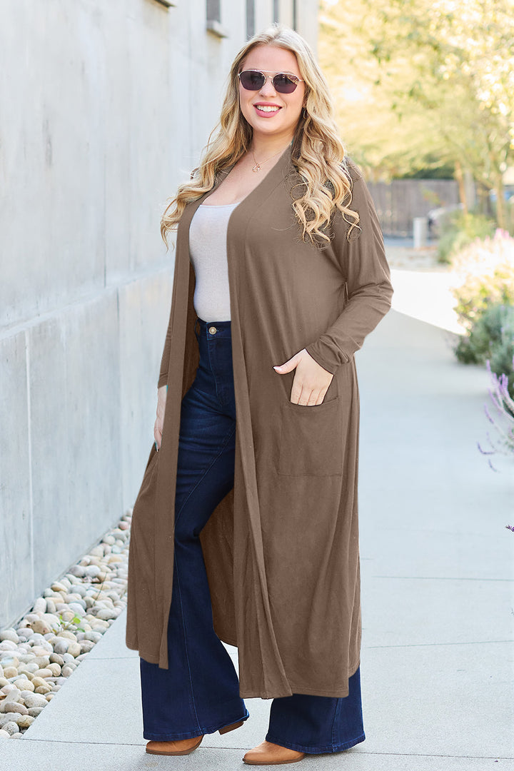 Wearing sunglasses, a woman with long blonde hair stands on a sidewalk bordered by rocks and plants, framed by buildings in the background. Her outfit features the Basic Bae Full Size Open Front Long Sleeve Cover Up in brown with its comfortable stretchy fabric, paired with a white top and dark flared jeans.