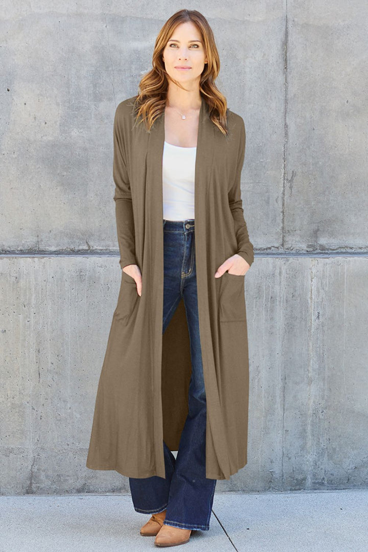 A person stands against a concrete wall wearing the Basic Bae Full Size Open Front Long Sleeve Cover Up, an olive-green cardigan made of stretchy material, paired with a white top and blue jeans. They have long hair and are wearing brown shoes. With their hands tucked into the cardigan pockets, they exude a relaxed demeanor.