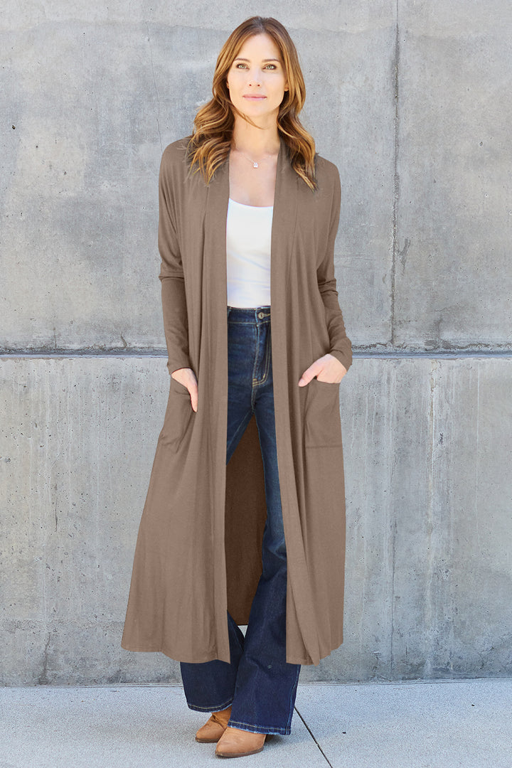 A woman stands against a concrete wall, wearing the Basic Bae Full Size Open Front Long Sleeve Cover Up made from stretchy material over a white top and dark blue jeans. She has her hands in the cover up pockets and is smiling. She has long, light brown hair and sports brown shoes.