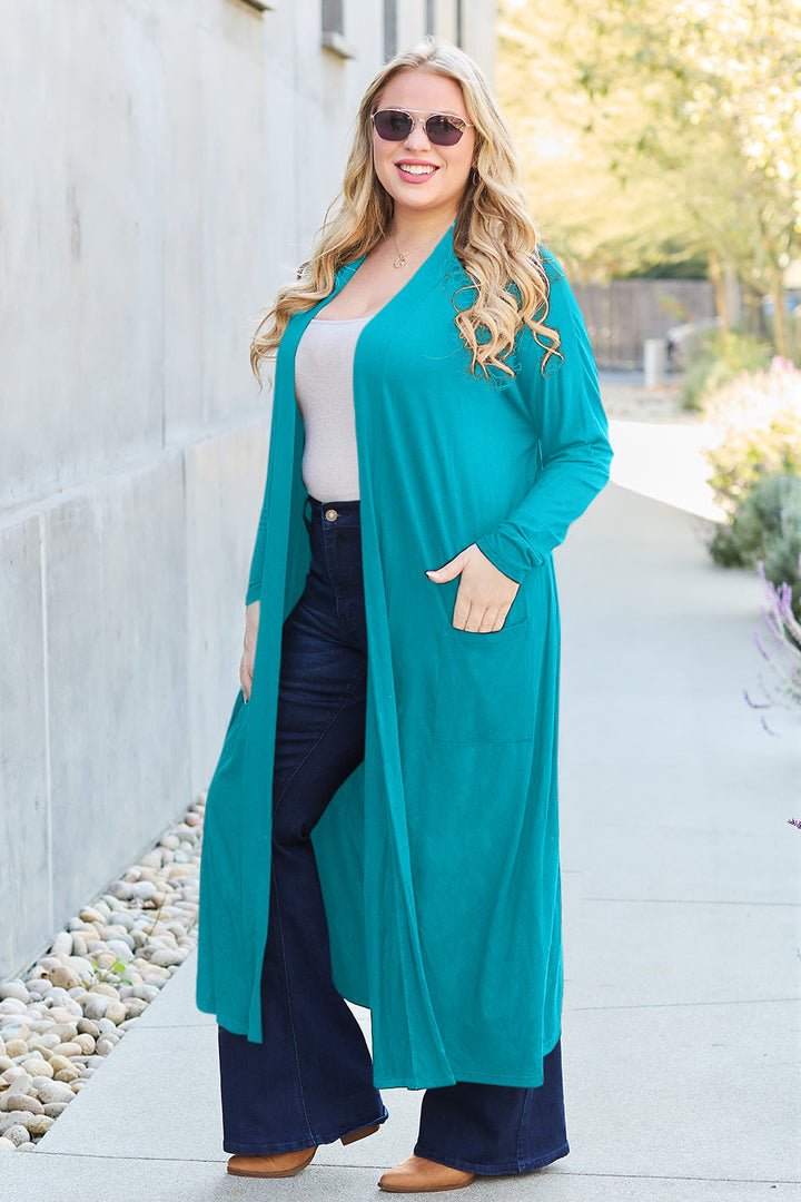 A woman with long blonde hair stands on a sunny sidewalk, wearing sunglasses and the Basic Bae Full Size Open Front Long Sleeve Cover Up in teal layered over a beige stretchy top. Her dark blue bell-bottom jeans add a stylish flair, while her brown shoes provide a grounded finish to the outfit. Greenery and a building serve as the backdrop.