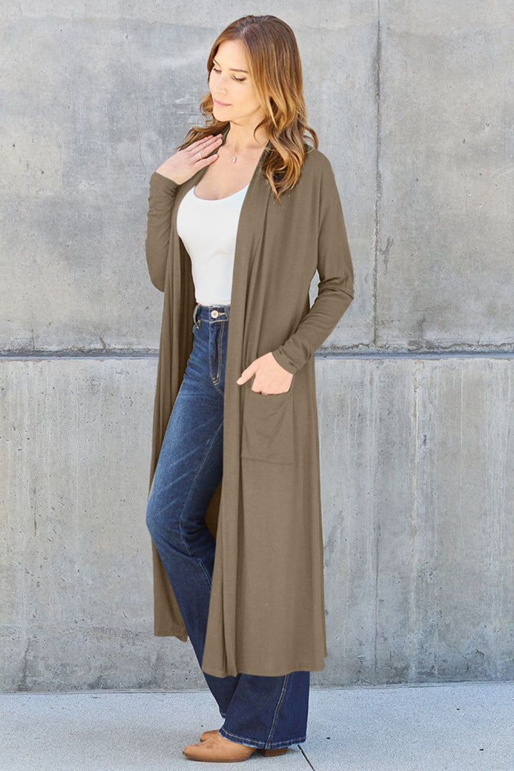A woman stands against a concrete wall, modeling the Basic Bae Full Size Open Front Long Sleeve Cover Up in a rich brown hue. She pairs it with a white top, blue jeans, and tan shoes. Her long brown hair cascades down as she gently touches her neck while gazing downward. The ensemble is ideal for those who enjoy garments that are easy to care for with machine wash cold.
