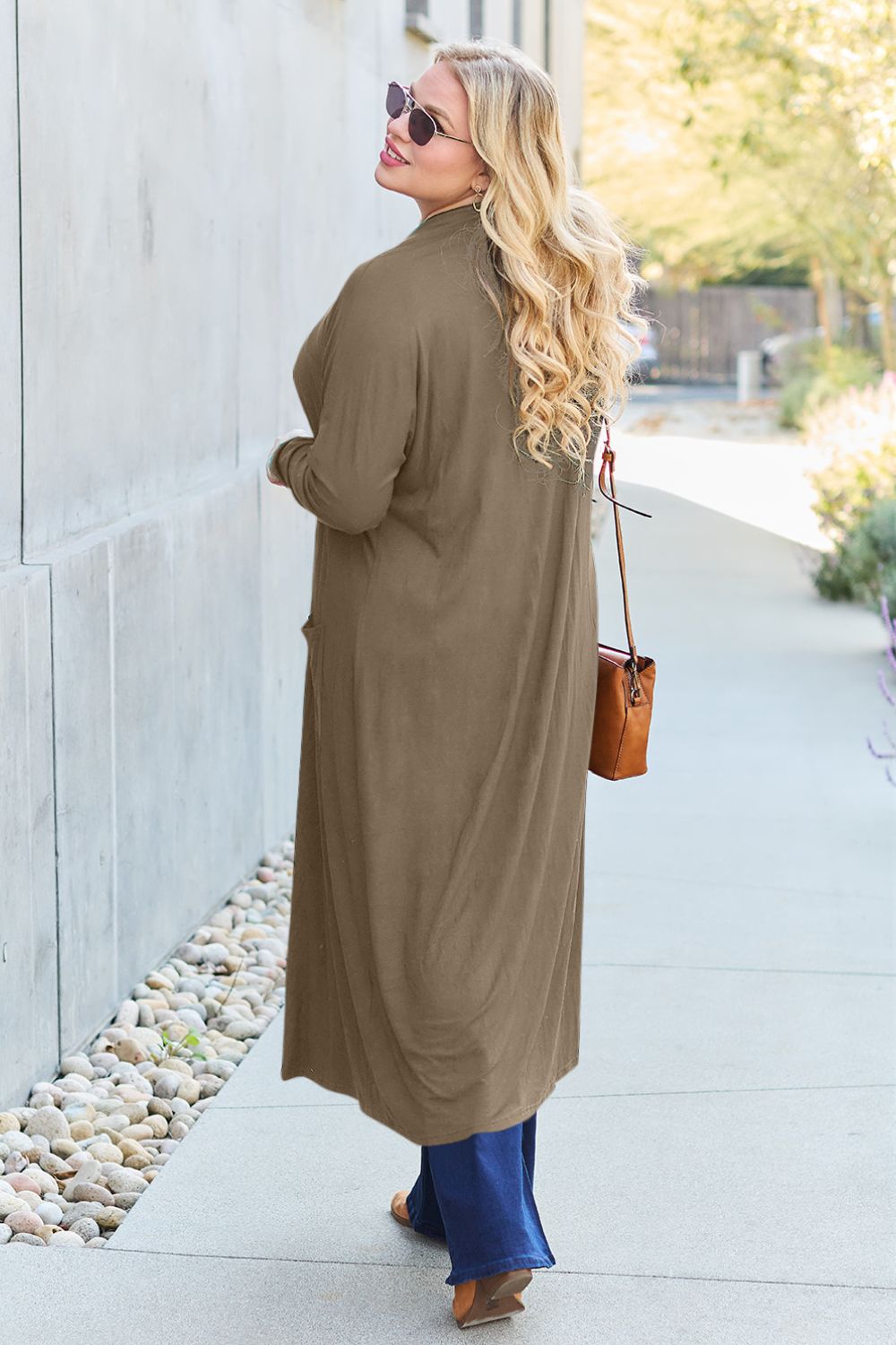 A woman with long blonde hair wearing sunglasses, a Basic Bae Full Size Open Front Long Sleeve Cover Up, blue jeans, and a brown purse walks along the sidewalk. There are stones beside the path and trees in the background.
