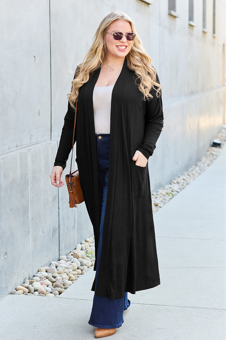 A person with long blonde hair and sunglasses is wearing the Basic Bae Full Size Open Front Long Sleeve Cover Up—a long black cardigan with pocketed sheer details—over a white top and blue jeans, while carrying a brown handbag as they walk along a modern gray building, appearing to smile.