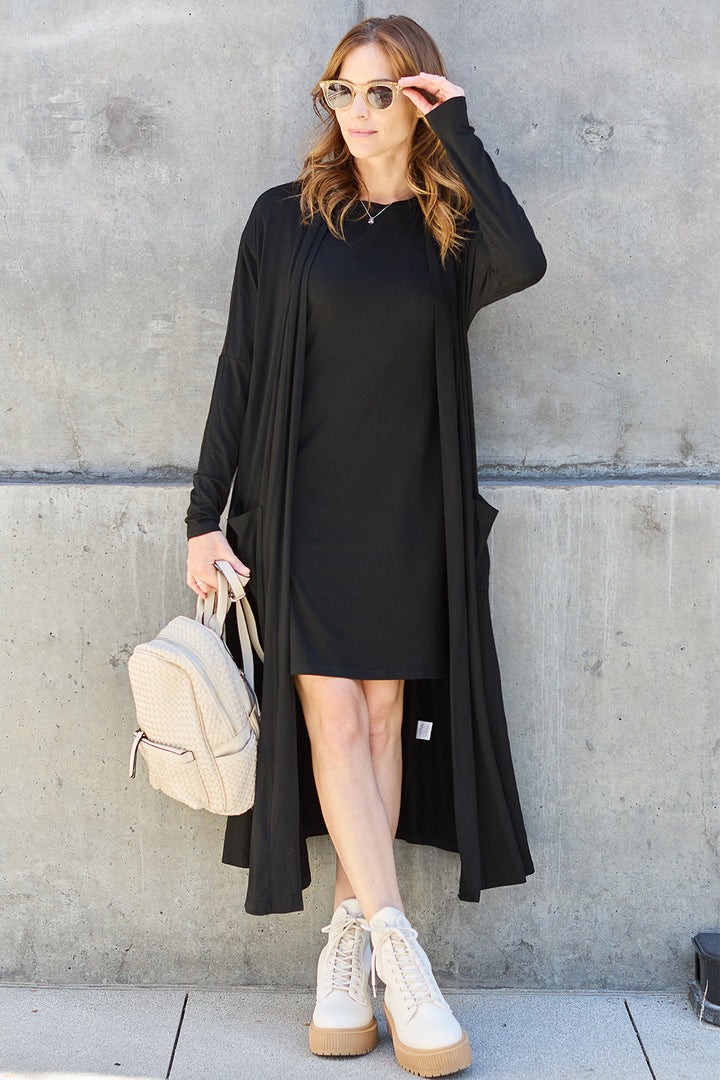 Dressed in the Basic Bae Full Size Open Front Long Sleeve Cover Up cardigan over a black dress, the individual stands against a concrete wall. Holding a light-colored backpack and sporting sunglasses along with beige boots, they adjust their shades with their right hand, adding an air of mystery to their pocketed sheer ensemble.